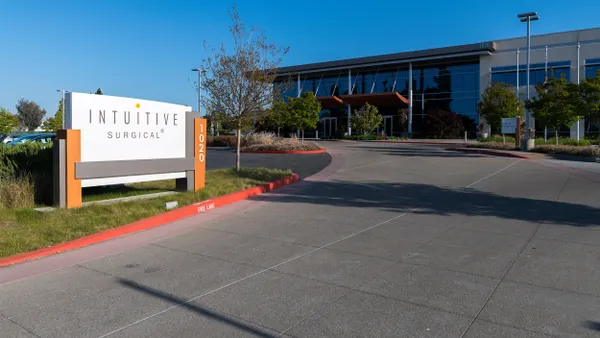 A modern office building with large reflective glass windows is shown behind a wide driveway and landscaped entrance. In the foreground, a white and orange monument sign reads "Intuitive Surgical" with the number "1020" vertically on the side.