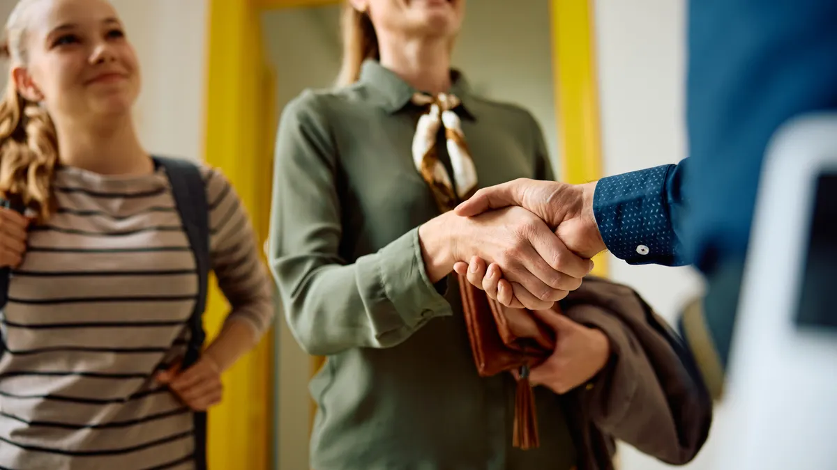 An adult is shown shaking hands with another adult while a student carrying a backpack is seen standing to the side. The hands are predominate in the photo.
