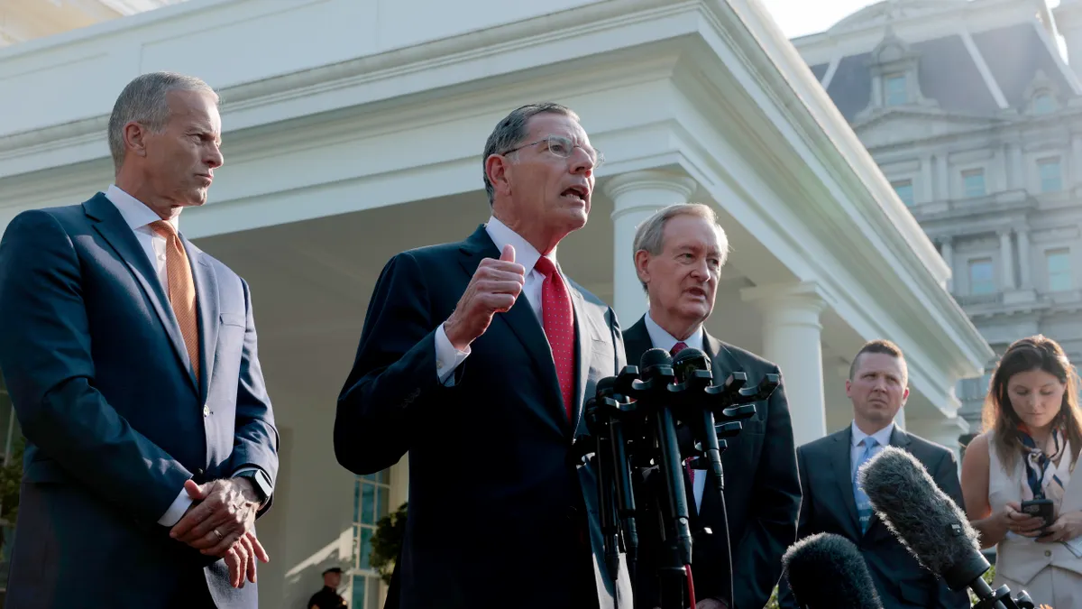A man wearing a suit and tie speaks into microphones while two men wearing suits and ties look on.