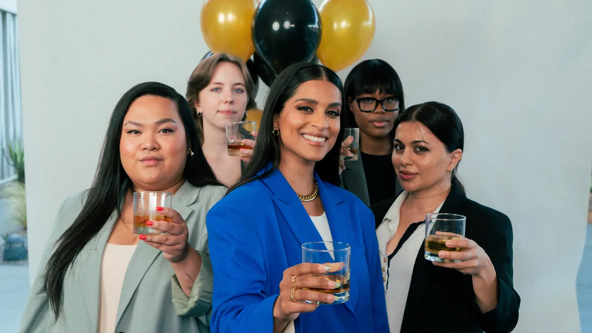 A group of diverse women hold up glasses of scotch in front of balloons.