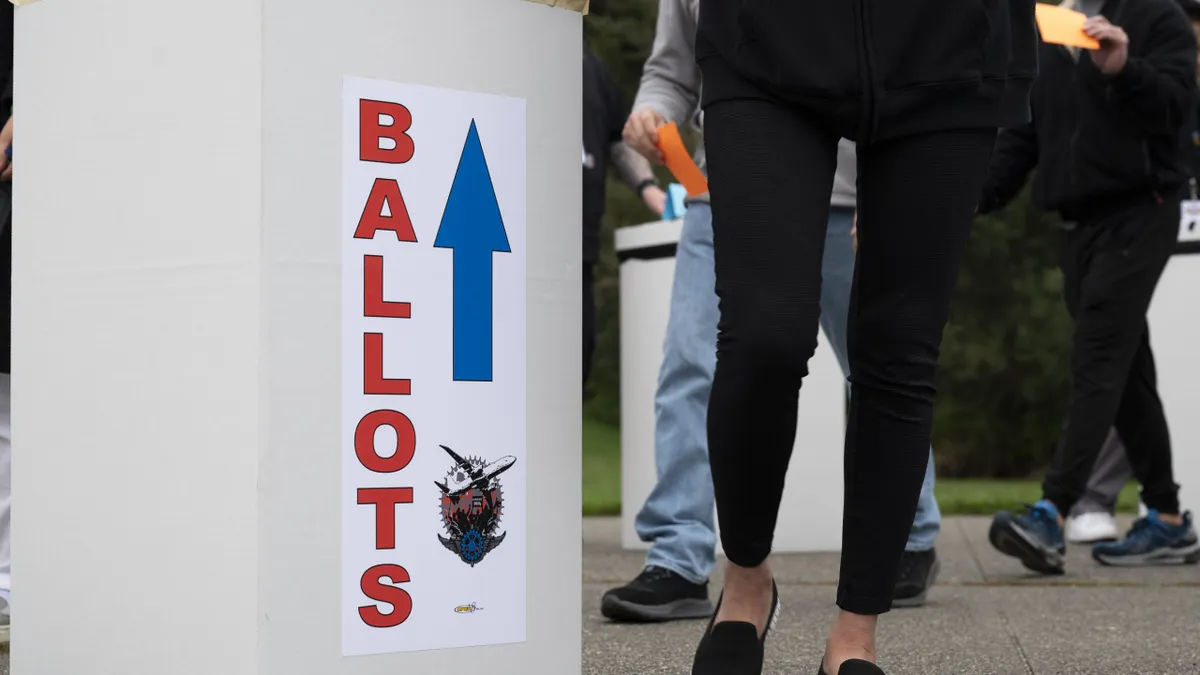 A person in black pants and shoes standing next to a light yellow box with "Ballot" in red text and a blue arrow pointing up.