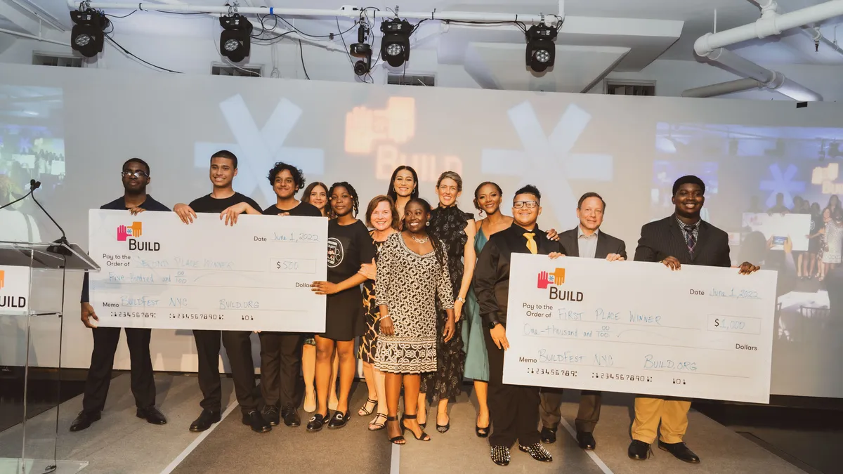 A group of students and adults stand looking at the camera and holding two large checks