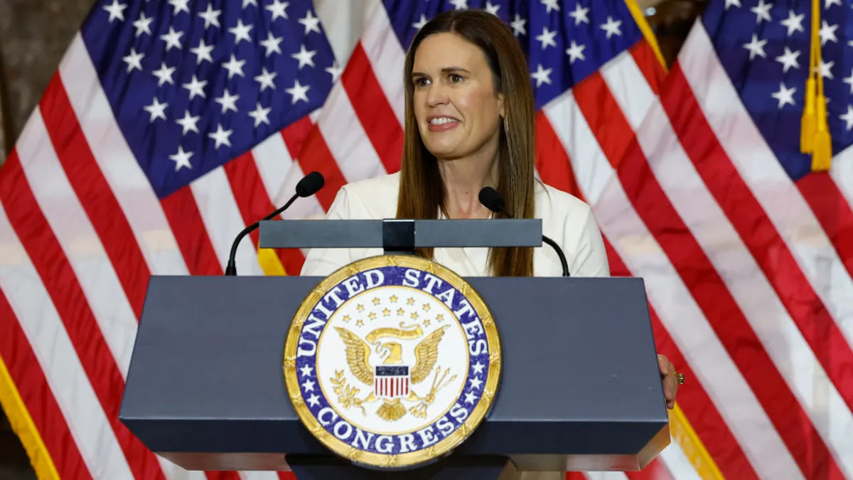 Sarah Sanders in white stands at podium with three American flags in the background
