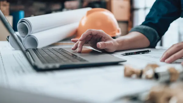 A person works at a laptop with a bundle of large, rolled-up blueprints on the side in the background.
