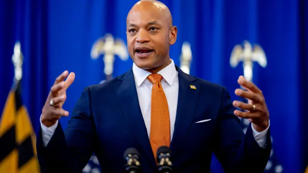 Gov. Wes Moore speaks while raising his hands at a podium in front of a blue background.