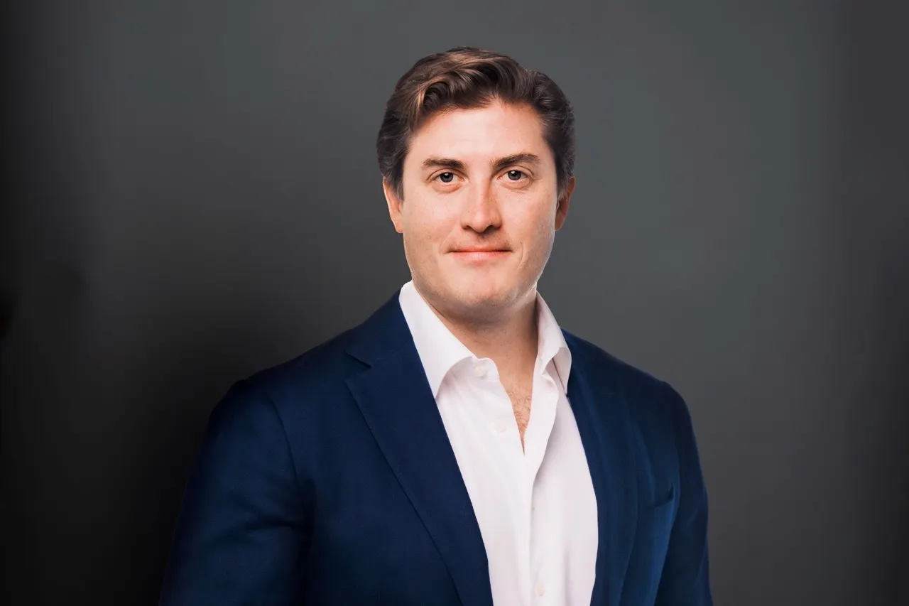 A head and shoulders shot of a person in a dark suit against a gray background.