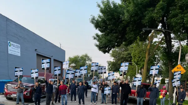 Two dozen people holding picket signs saying they're striking against Mauser Packaging Solutions stand outside an industrial facility.
