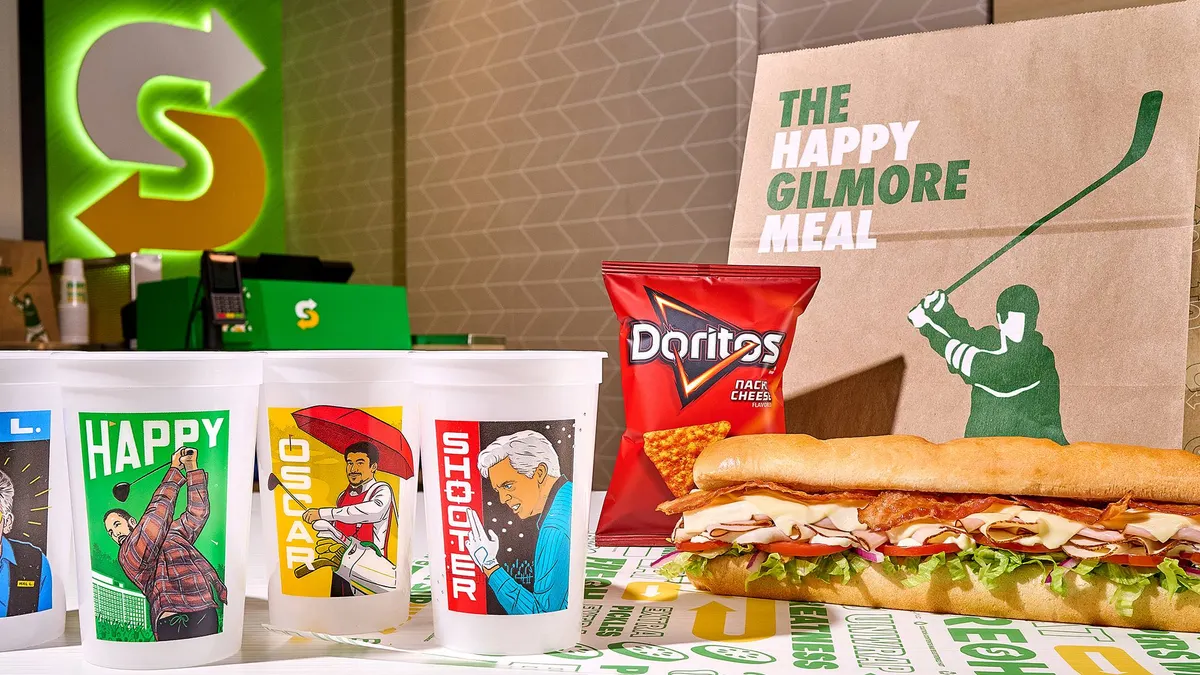 A row of four cups with illustrations of "Happy Gilmore 2" characters are lined up next to a Subway sandwich.