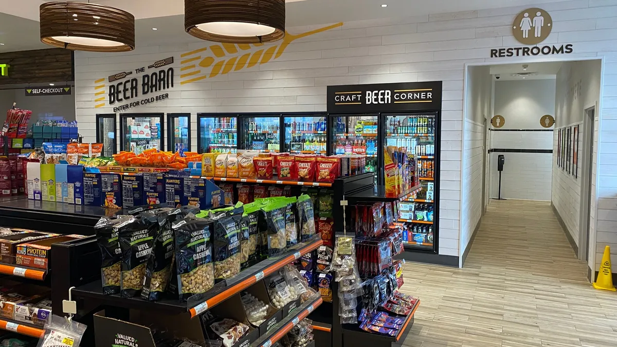 A photo of the interior of a convenience store, showing shelves full of snacks and a beer cave set in the back wall. Signs over the beer gave say "the beer barn. enter for cold beer." while a sign over a hallway to the right of the beer cave says "restrooms."