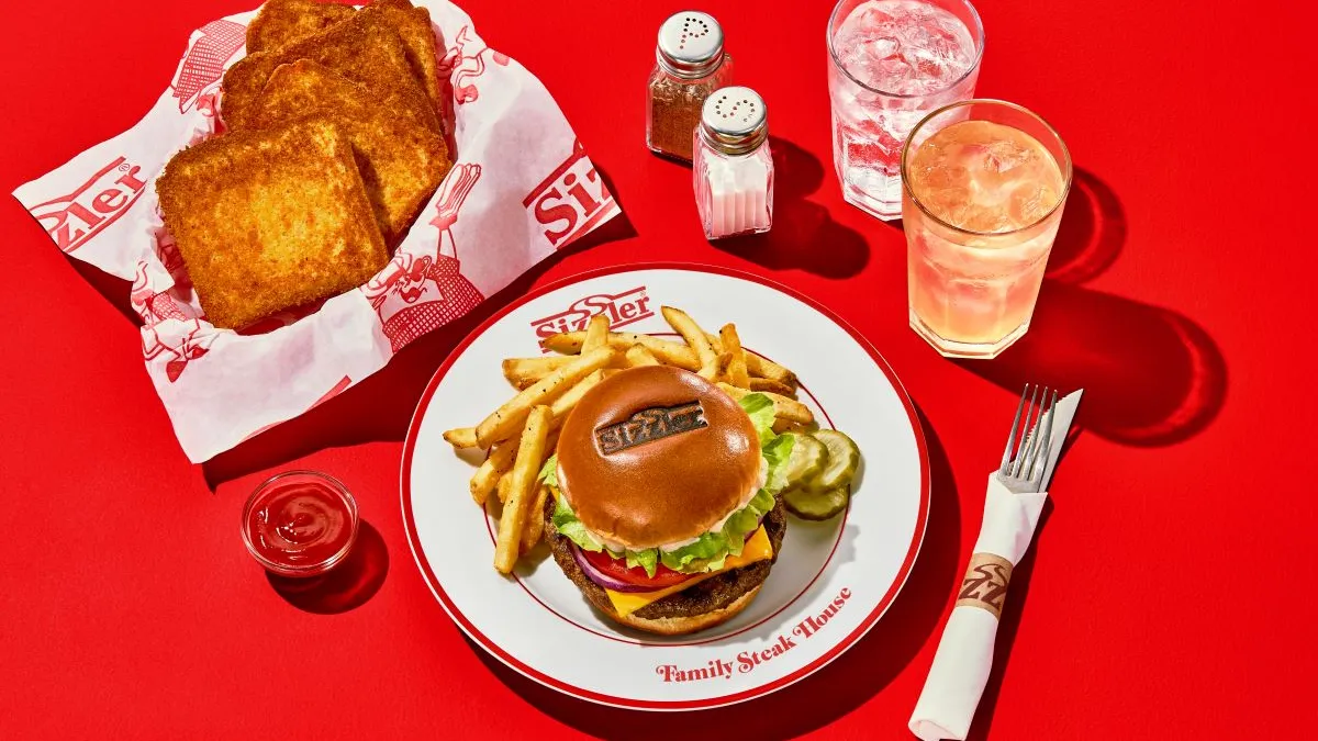 A white plate with the words "sizzler" and "family steakhouse" with a burger, fries and pickles. The photo also includes a basket of cheese toast, wrapped silverware and two drinks.