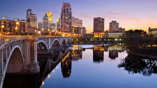 An image of the Minneapolis downtown skyline at sunset, with a bridge over a river reflecting tall buildings against a dark blue sky.