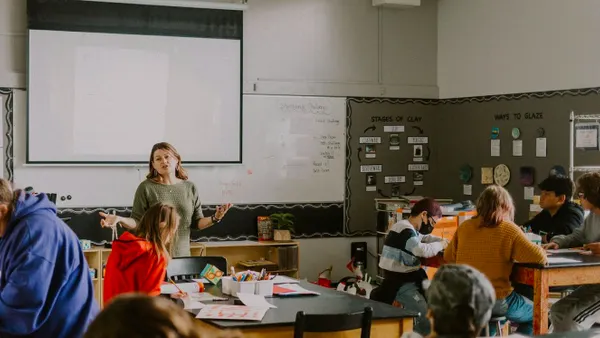 Students are sitting at desks in a classroom. An adult is standing.