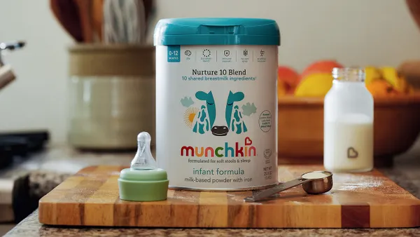 A container of Munchkin-branded infant formula is displayed on a cutting board next to a bottle of milk, a bottle nipple and a measuring spoon.