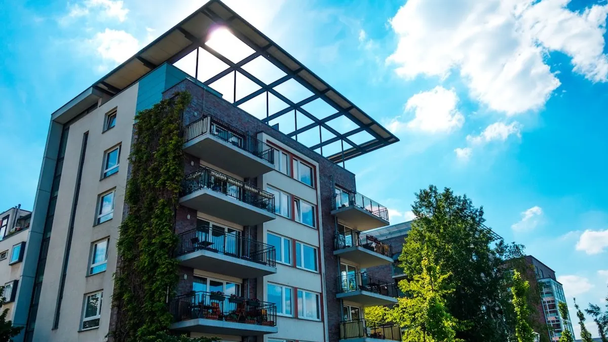 A modern apartment building under a clear blue sky with scattered clouds. Greenery climbs one side, enhancing the contemporary, urban aesthetic.