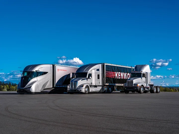 Kenworth's SuperTruck 2, T680 and T600A trucks lined up in a parking area in front of blue skies with clouds.