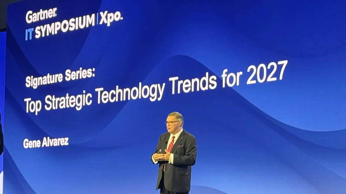 An image of Gene Alvarez in a suit speaking onstage at the Gartner IT Symposium, with a blue background.