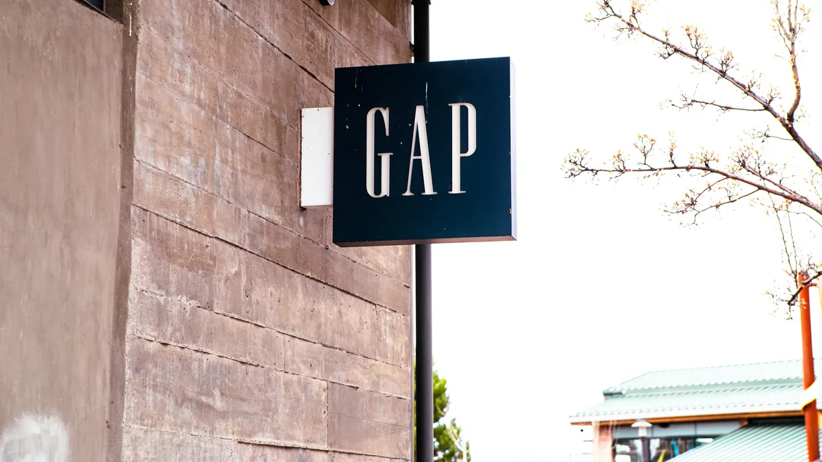 Branches with small buds jutting from the right point toward a blue Gap sign with white lettering, mounted on a beige stone building.