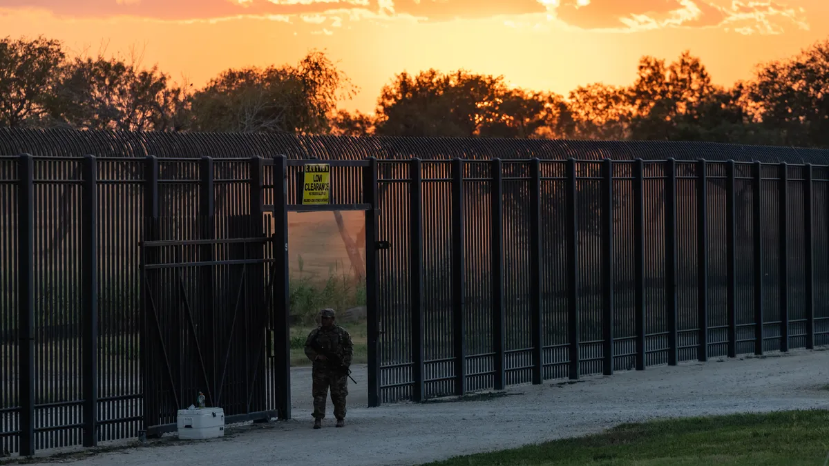 A person in uniform stands with a gun in front of a tall black fence as the sun sets in the background.