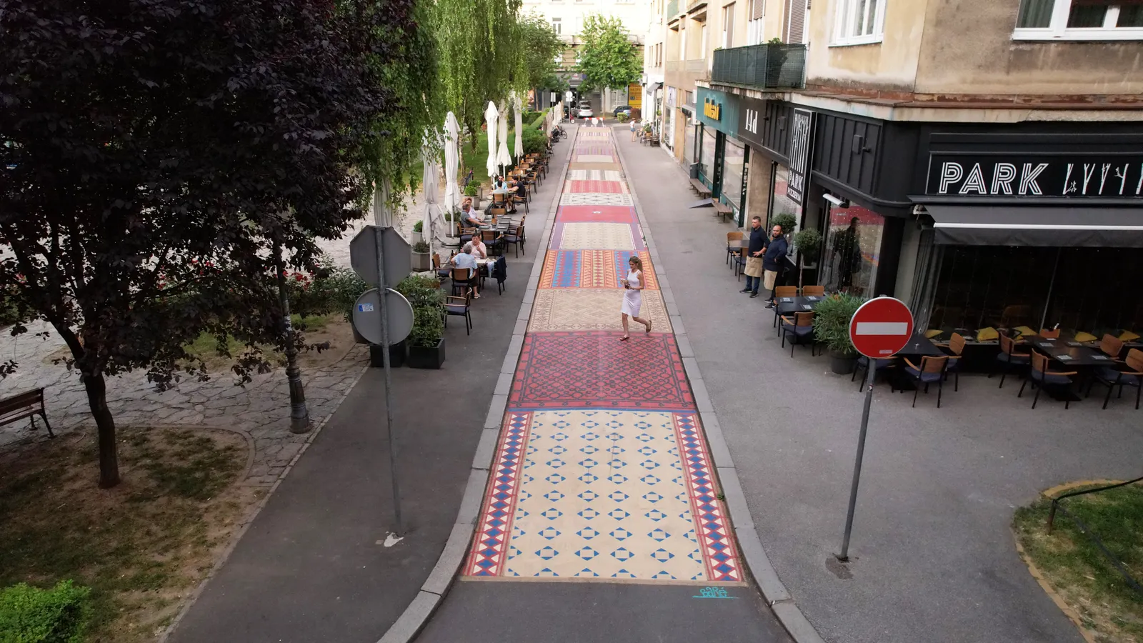 A person walks across a street painted with a patterned mural. Trees line one side of the street, and buildings line the other.