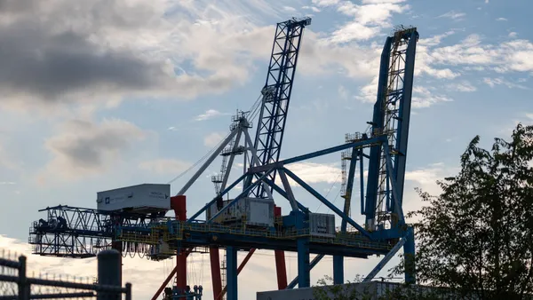 Large cranes and other machinery loom in the background in the dusk. Some greenery exists next to it.