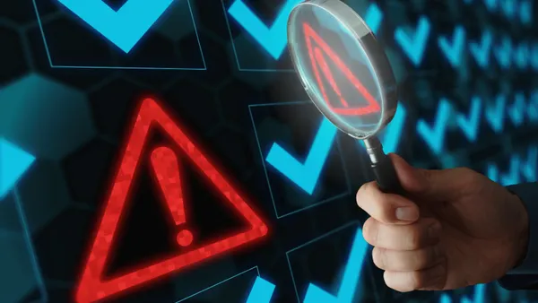 A hand holds a magnifying glass up to a red exclamation point enclosed by a red triangle