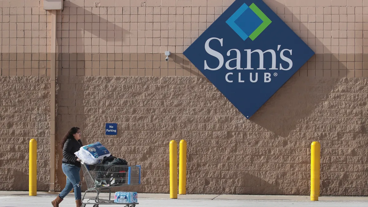 A shopper with a shopping cart full of merchandise stands in front of a Sam's Club sign outside the store