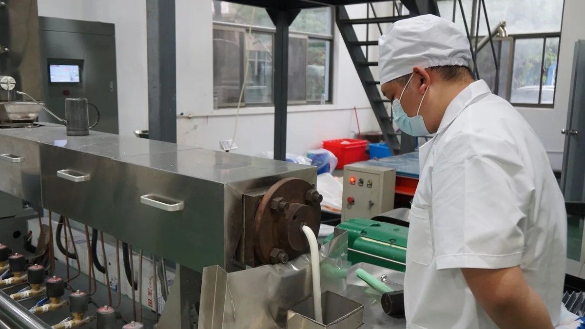 A person wearing a white shirt, cap and face mask stands next to industrial equipment that is producing a white material.