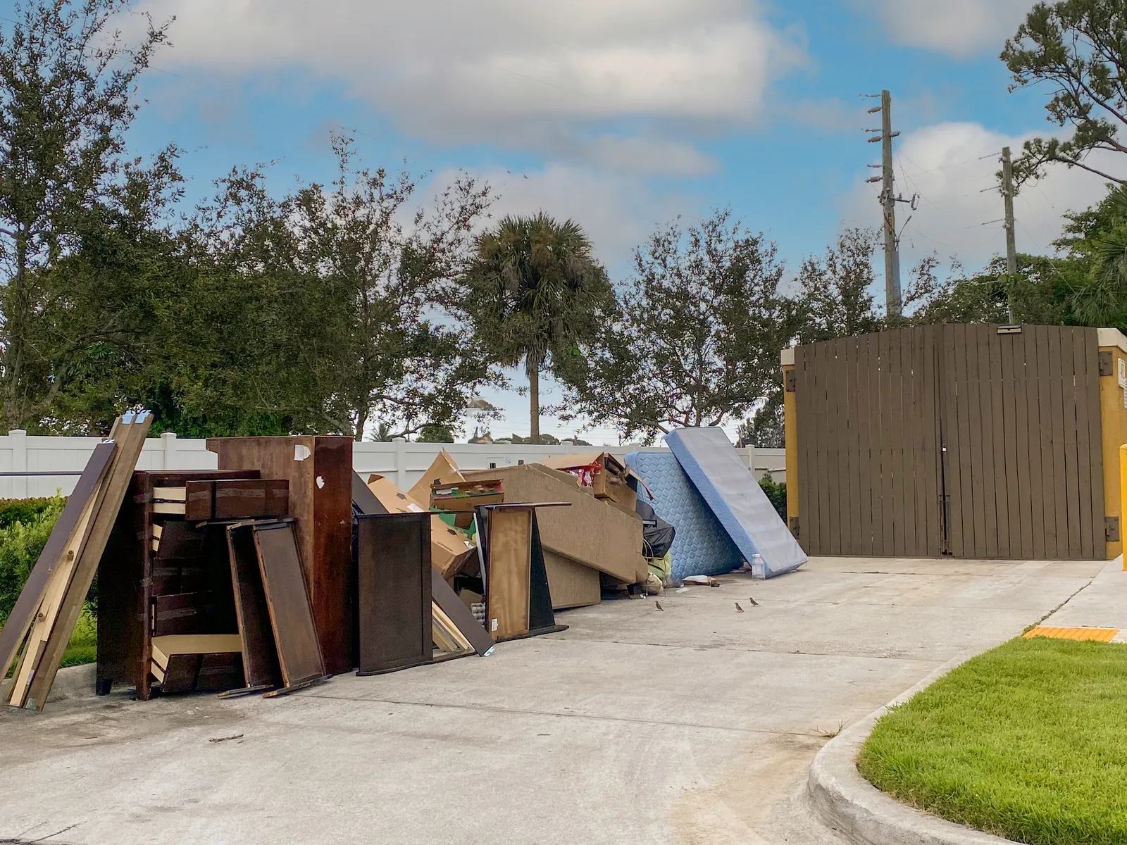 A stack of furniture and mattresses next to a trash enclosure at a multifamily building, with trees in the background.