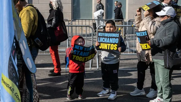 Two children stand in the middle of the photo, holding signs that read "New Yorkers fight together" in English and in Spanish. They are standing outside on a street and adults are also present holding signs.