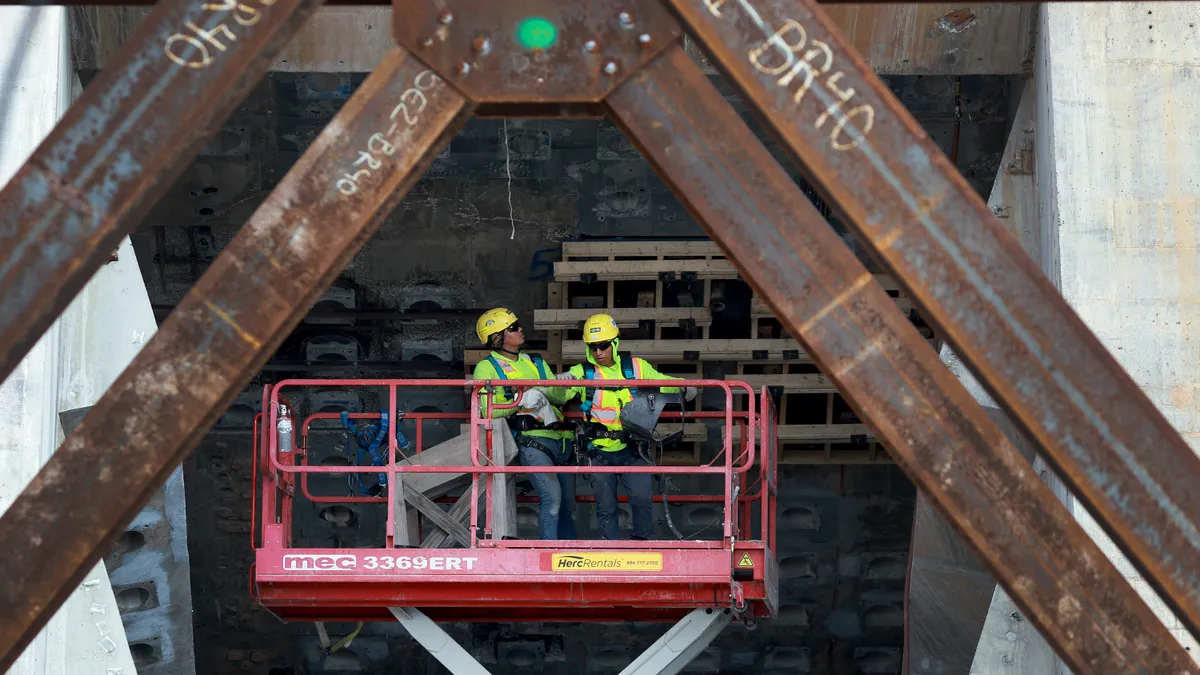 Two construction workers ride a lift underneath a rusty bridge.