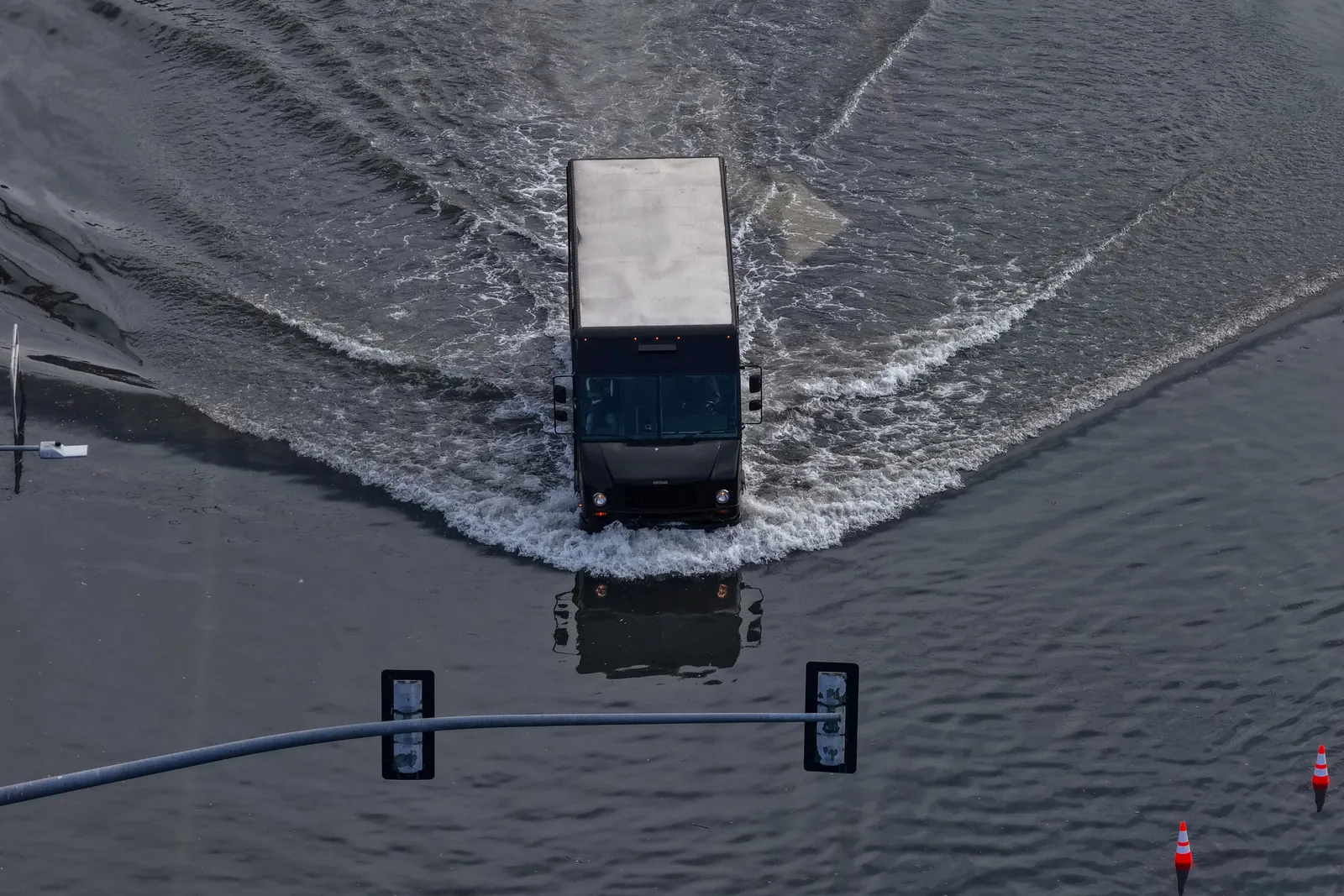 A big dark colored van drives through a flooded street, leaving ripples in its wake. Orange cones stand in the right corner while a traffic light stretches across the bottom of the image.