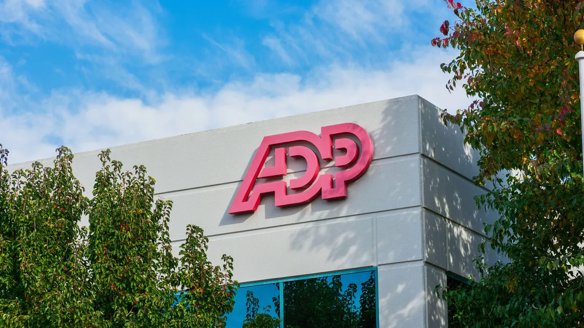 White building with the ADP logo in bold red letters near the roof, partially obscured by leafy trees and under a blue sky with scattered clouds.
