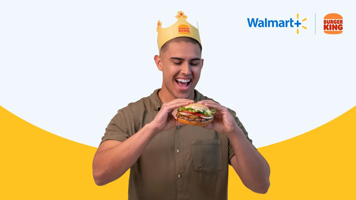 An image of a man wearing a Burger King crown and holding a Whopper. He is about to eat a Whopper and has his mouth open wide.