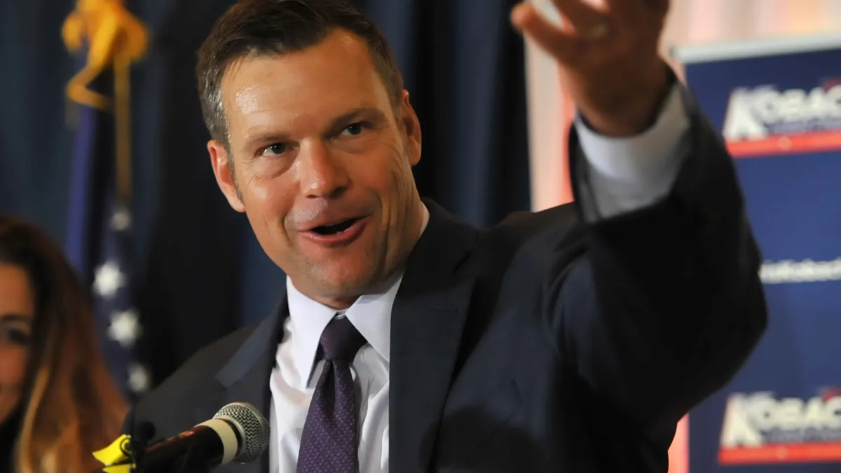 A White man speaks at a podium, gesturing with his left hand