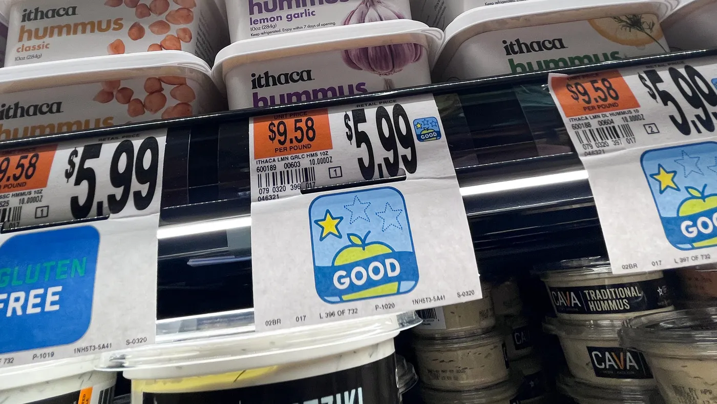 Price tags on a supermarket shelf that read “$5.99” and either “gluten free” or “good” beneath containers of hummus.