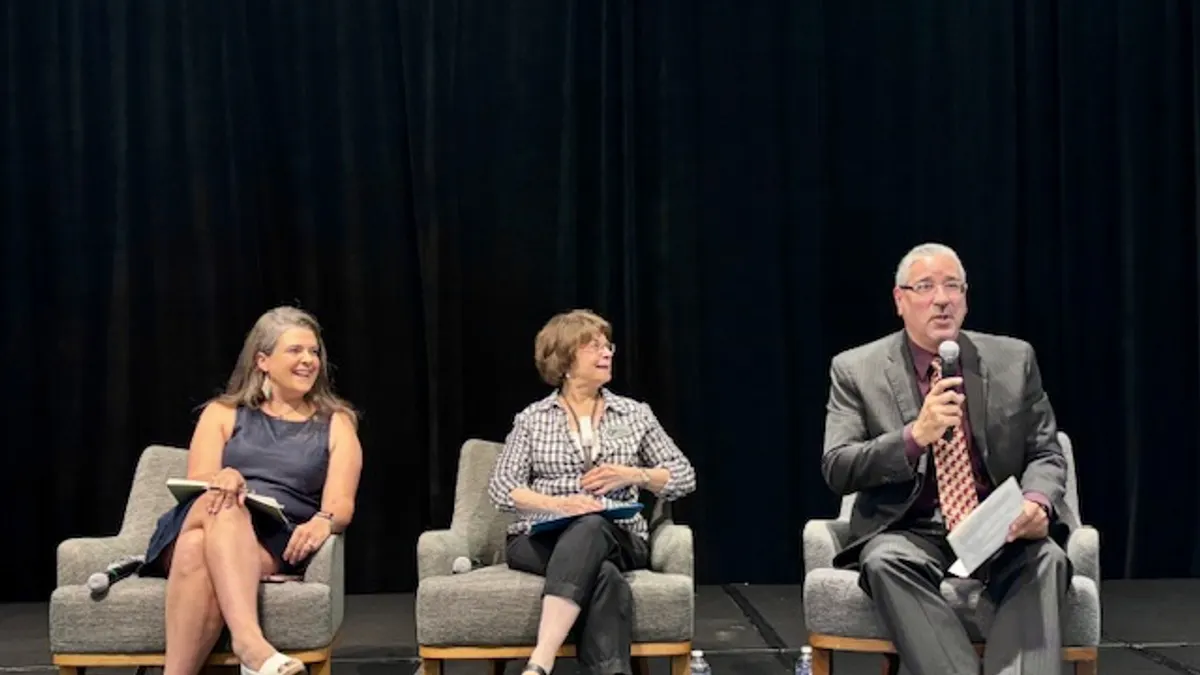 Three people are sitting in chairs on a stage. One person has a microphone