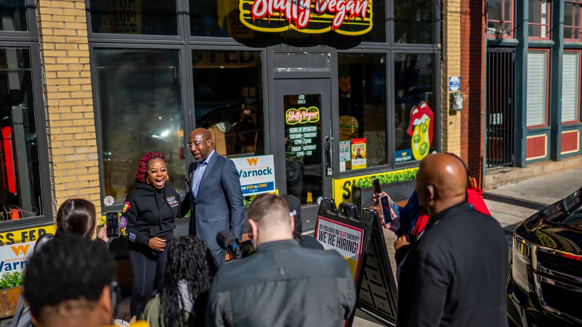 Slutty Vegan founder and CEO Pinky Cole and Senator Raphael Warnock stand in front of a Slutty Vegan restaurant, surrounded by press.