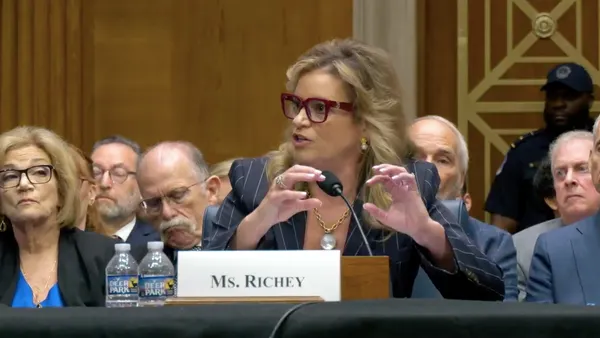 Kimberly Richey speaks into a microphone, sitting behind a placard bearing her name