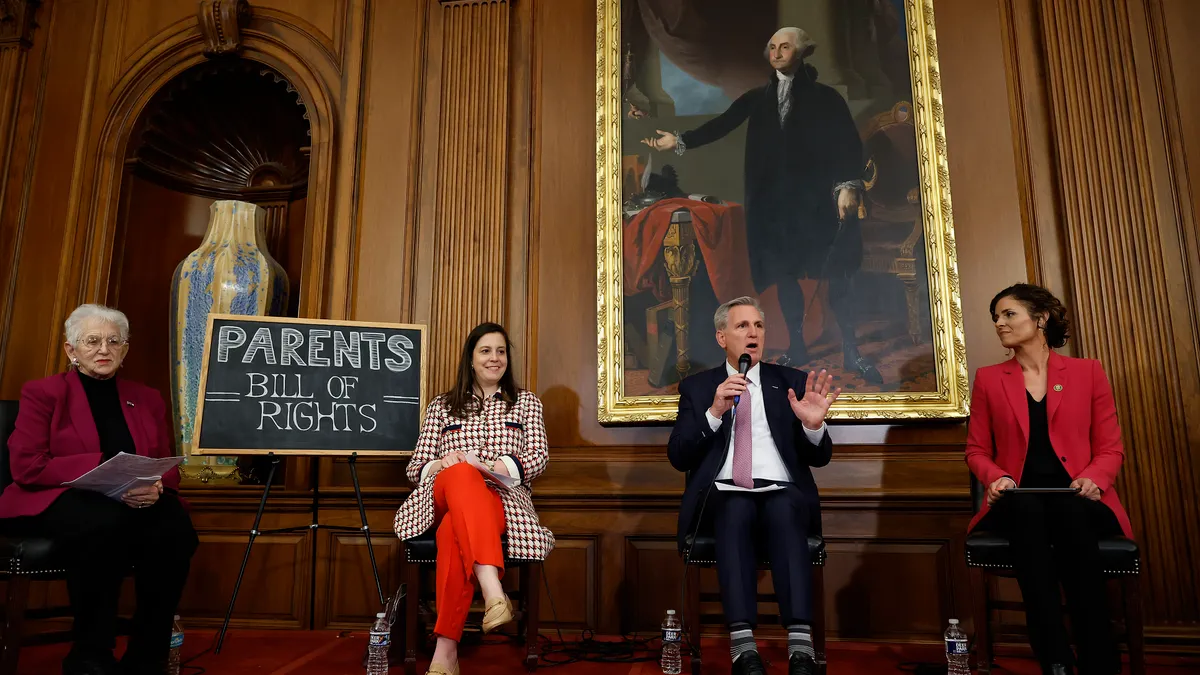 Four adults are seated on a stage in a wood-paneled room. Next to them is a stand with a chalkboard sign that reads "Parents Bill Of Rights."