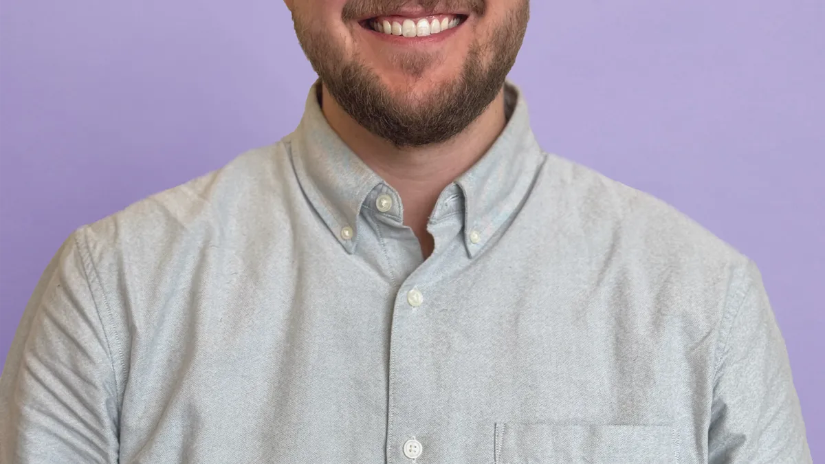 A headshot of a man in a blue button-down shirt against a purple background