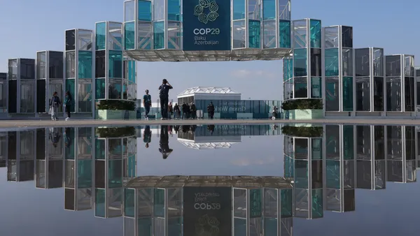 A semi-transparent arch to an event venue is reflected in a puddle. The arch is emblazoned with a sign reading "COP29, Baku, Azerbaijan."