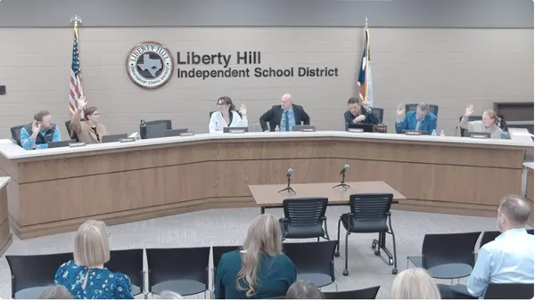 Seven people sit behind a dias and six of them are raising their hands. Behind them is a sign reading Liberty Hill Independent School District.