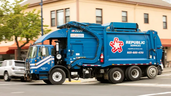 A blue Republic Services garbage truck is driving on a city street past beige buildings and storefronts. The side of the truck features a large white and red star logo with text. The truck also displays the number 2237 and “Clean Air Vehicle.”