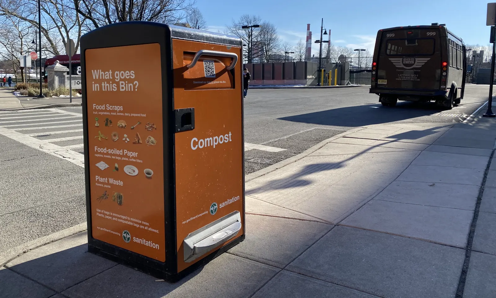 An orange bin with a handle to open labeled "Compost" sits on a sidewalk near a road.
