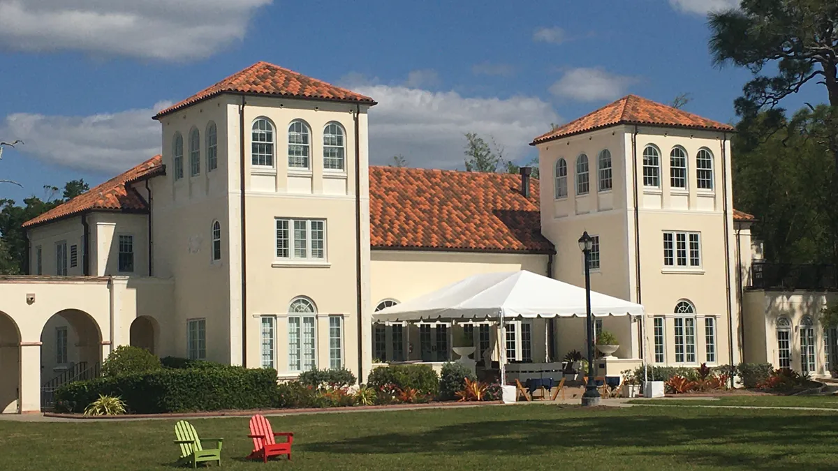 Two colorful lawn chairs sit on a lawn in front of  a large Spanish-style building.