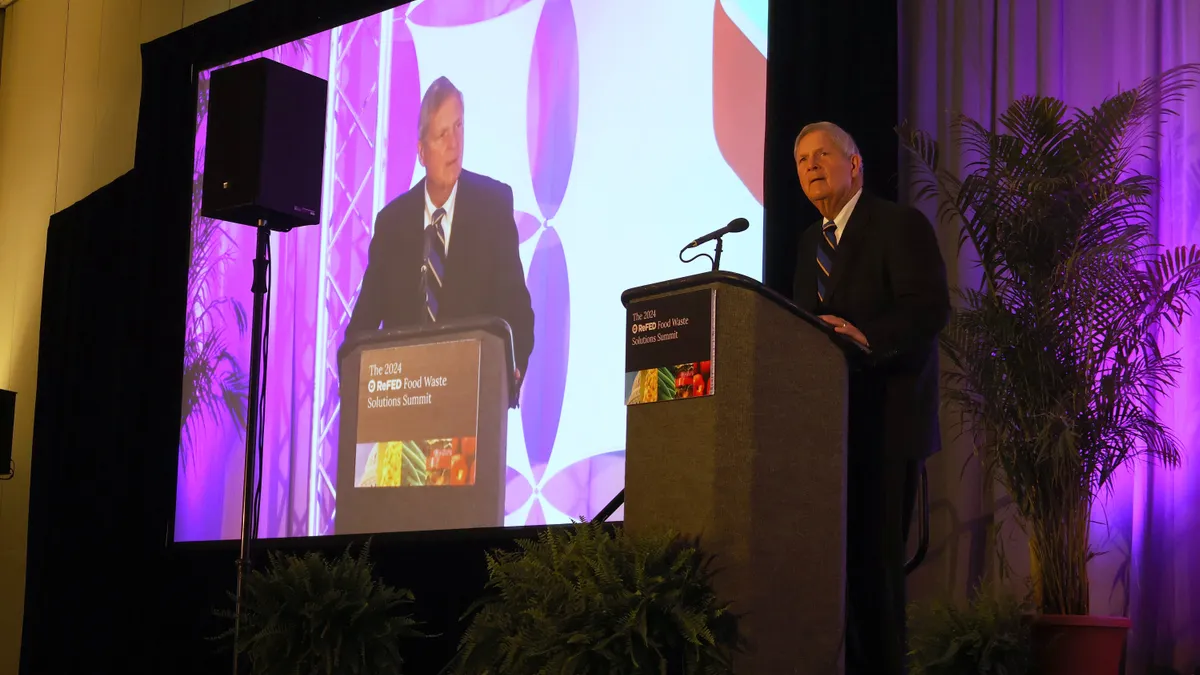 A man in a suit stands at a podium with a sign that reads "The 2024 ReFED Food Waste Solutions Summit." His image is projected on a screen behind him.