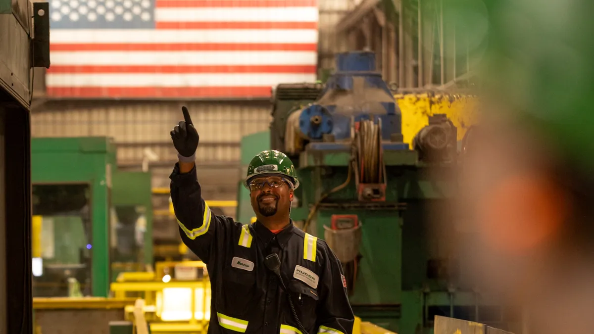 A person wearing a black and reflective yellow protective suit, gloves, hard hat and goggles, pointing up with a U.S. flag behind them.