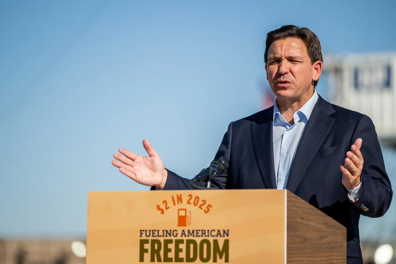 Ron DeSantis speaks in a navy blue suit and light blue shirt, at a wooden podium that reads "$2 in 2025" in red about a red gas can over the words "Fueling American Freedom" in blue.