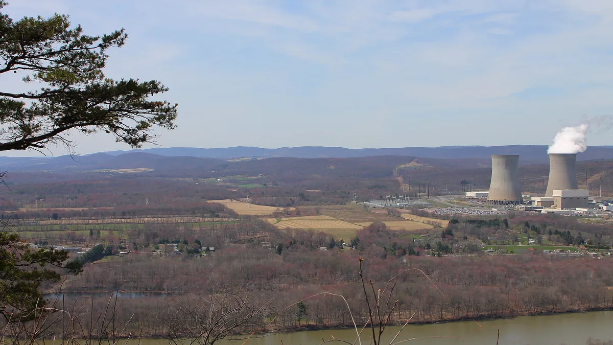 A river runs in front of forests, fields and steam billowing from two cement cooling towers from a power plant.