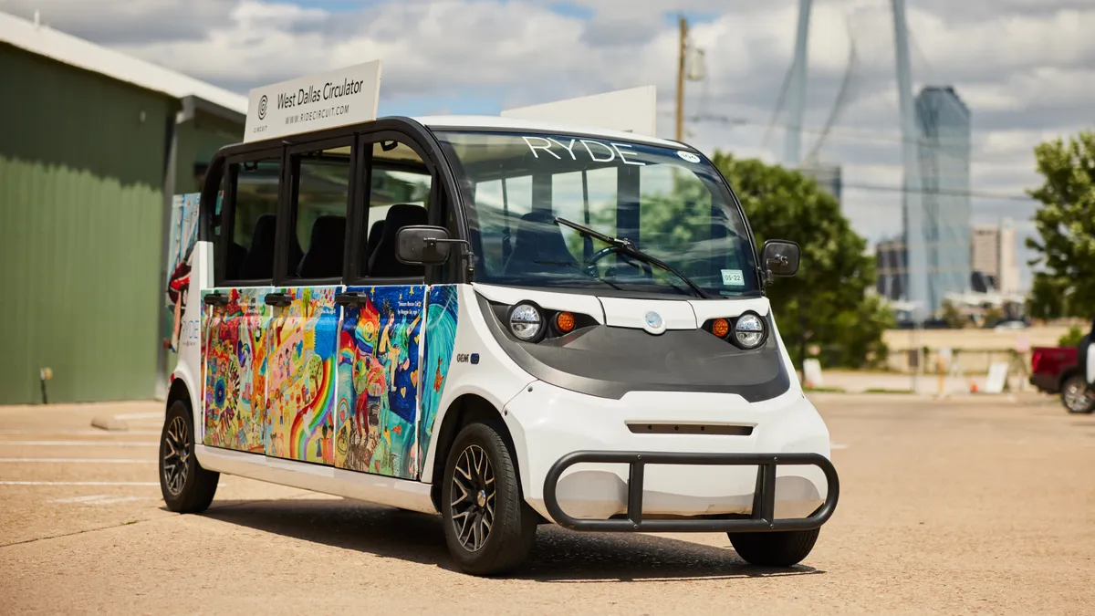 A four-wheel electric shuttle vehicle is parked in an industrial lot. A sign on the top side reads, "West Dallas Circulator."